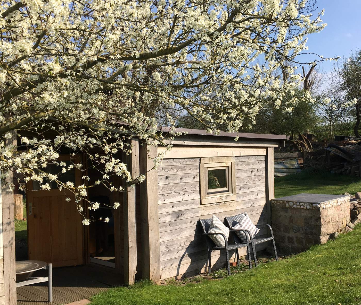 Das Blockhaus aus verwittertem Holz unter einem blühenden Obstbaum, rechts die gemauerte Grillstelle