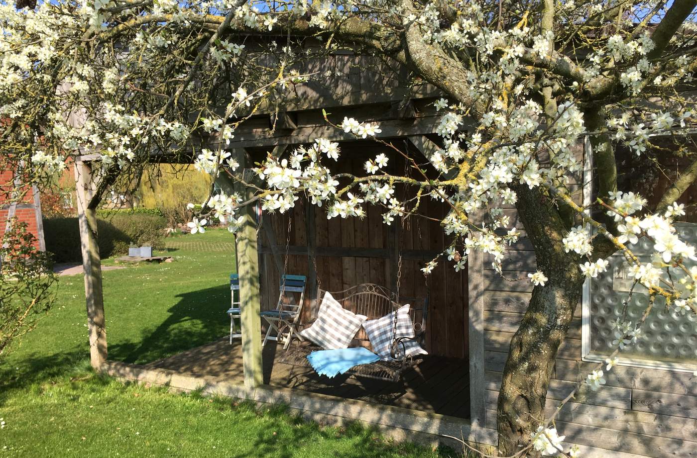 Schaukelbank mit Kissen unter dem überdachten Vorbau, davor ein blühender Baum